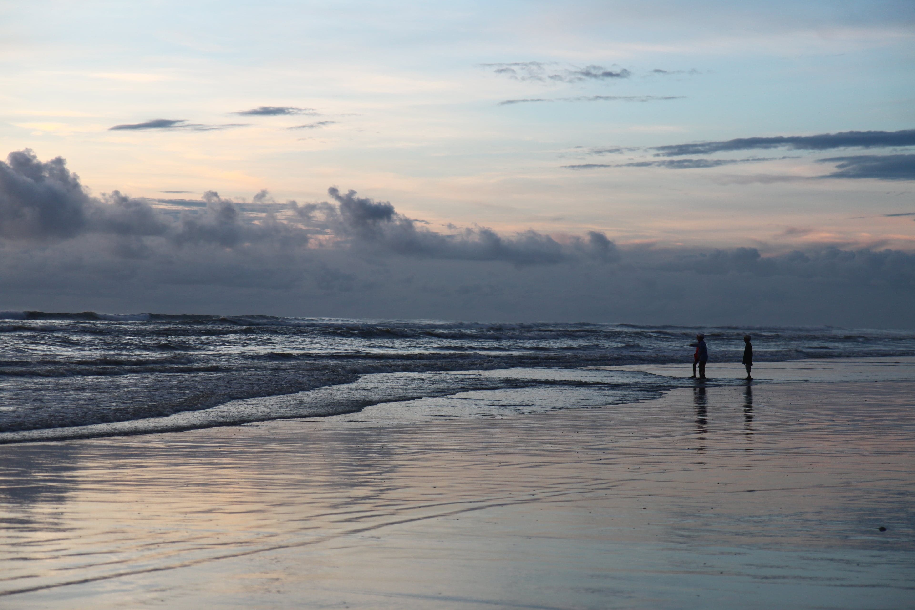 Two figures walking on the wet sand at Cox's Bazar beach at dusk, with a pink-and-grey sky above the sea