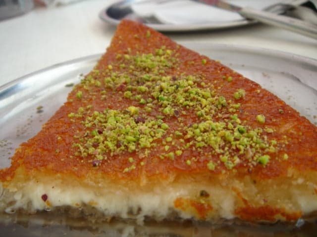 A slice of golden knafeh dripping with syrup and pistachios