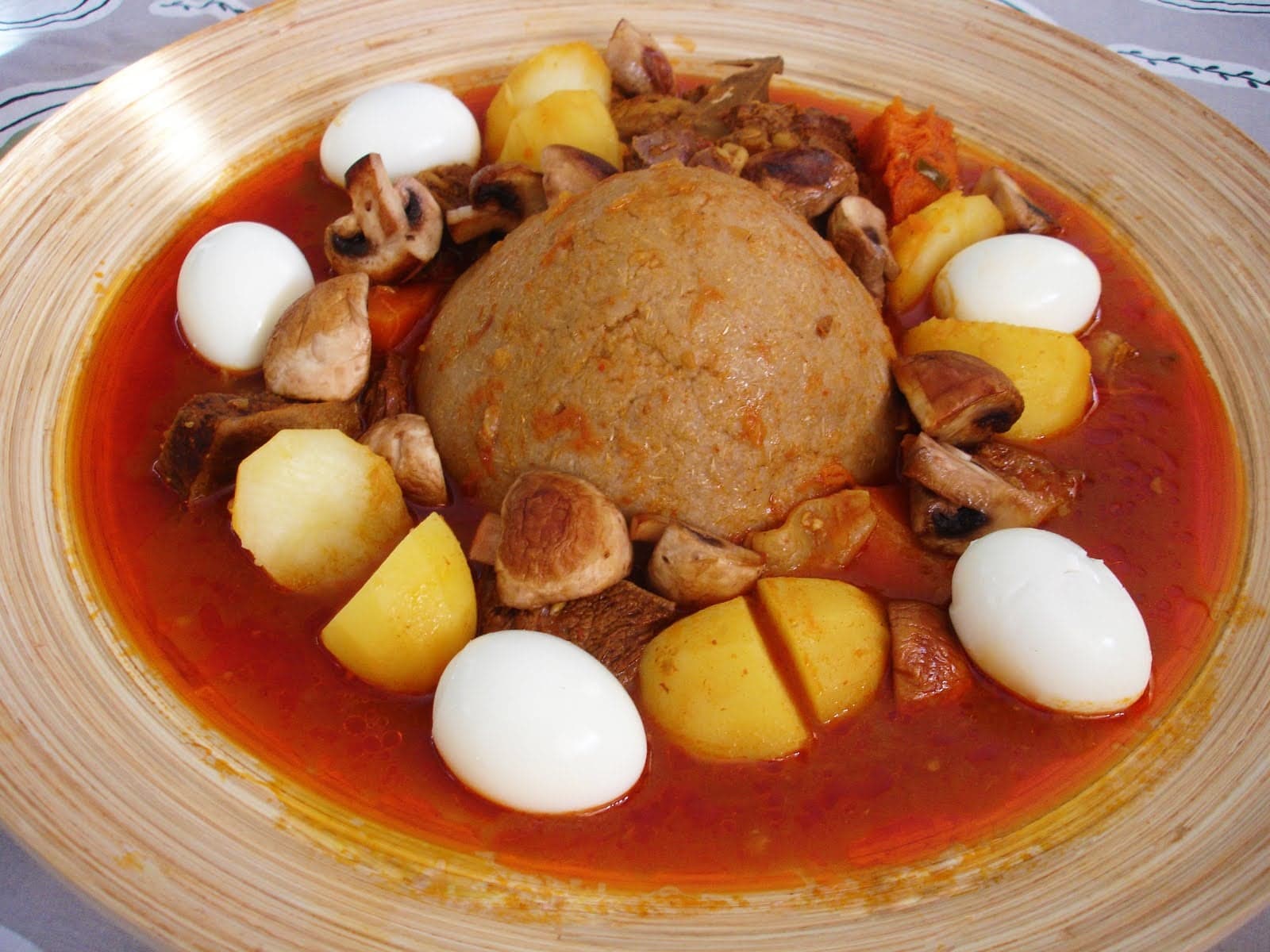 A smooth dome of bazeen dough surrounded by red lamb stew and hard-boiled eggs in a large dish