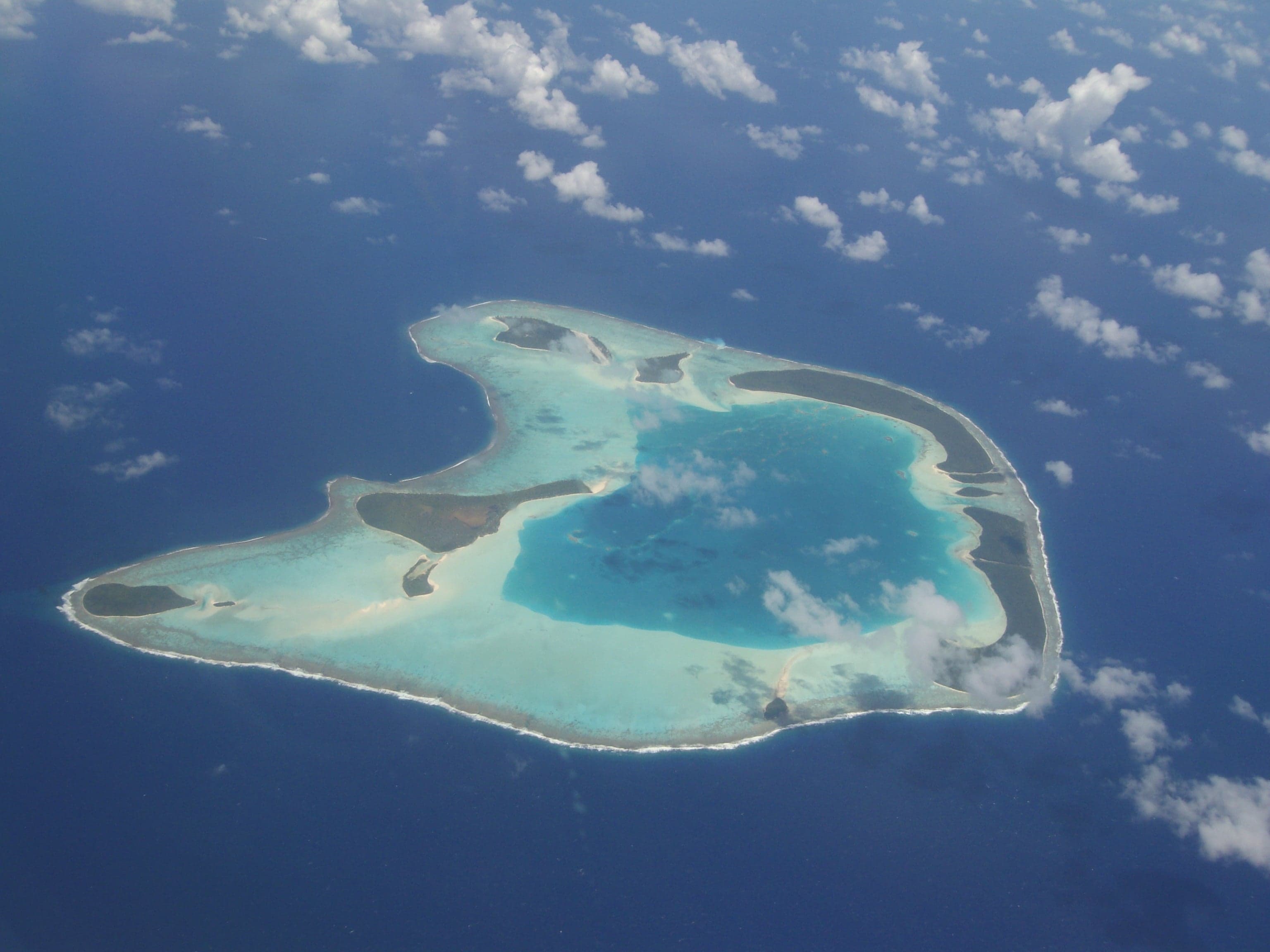 An aerial view of a green ring-shaped coral atoll surrounded by turquoise lagoon water and open ocean