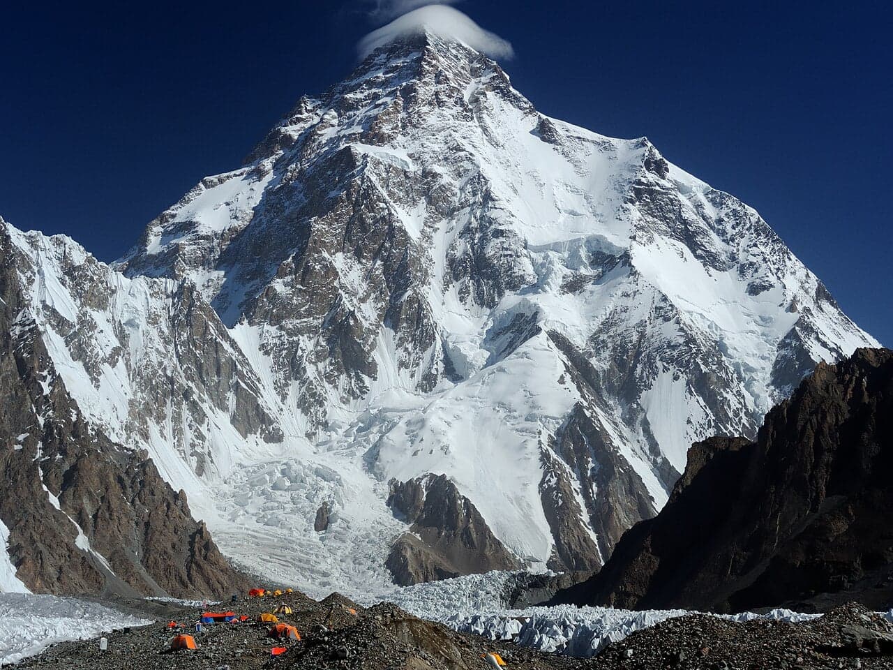 The pyramid-shaped peak of K2 rising above a glacier, with tiny orange tents at base camp