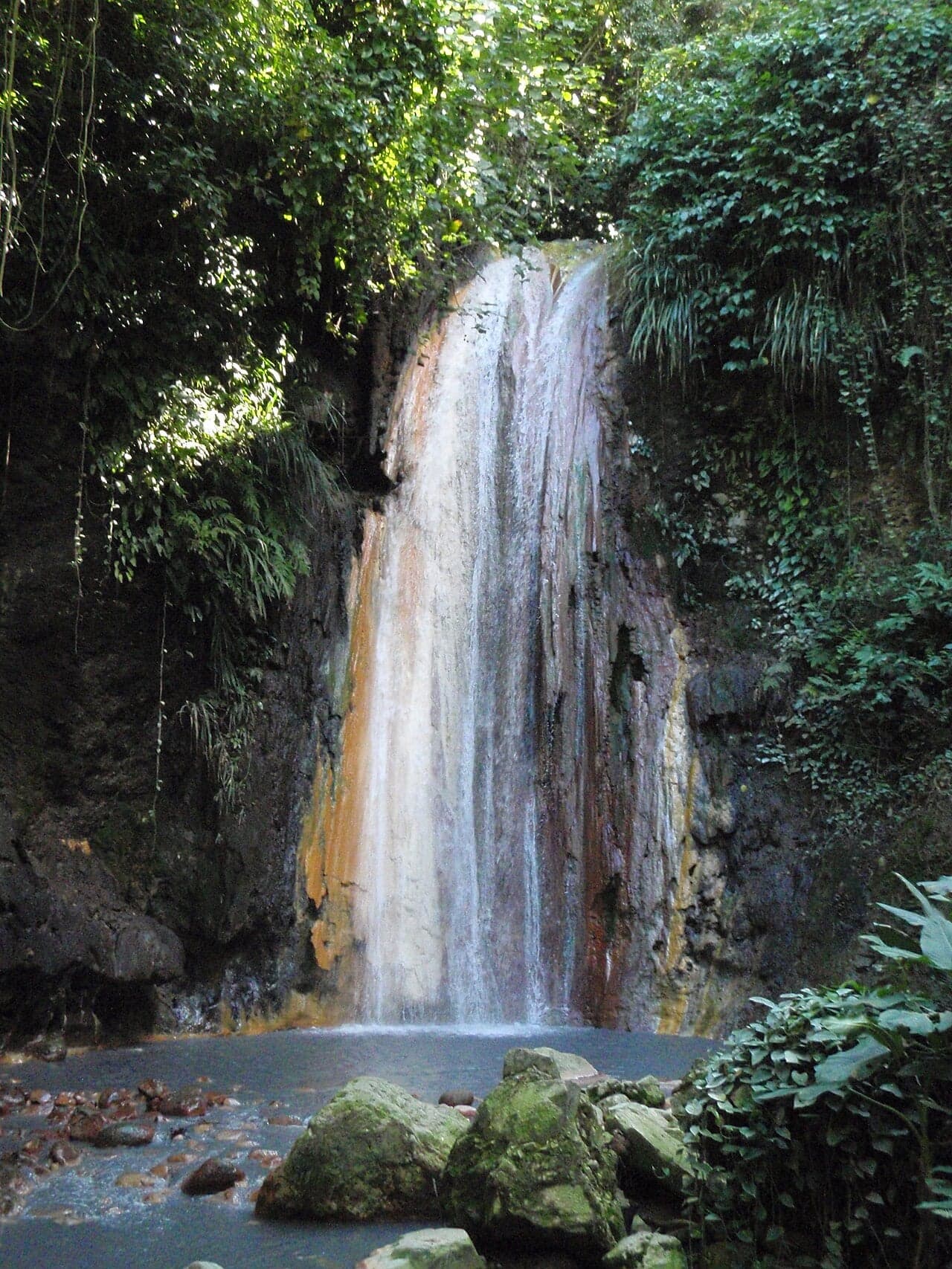 A waterfall with orange, yellow and purple mineral colours flowing into a pool amid tropical plants