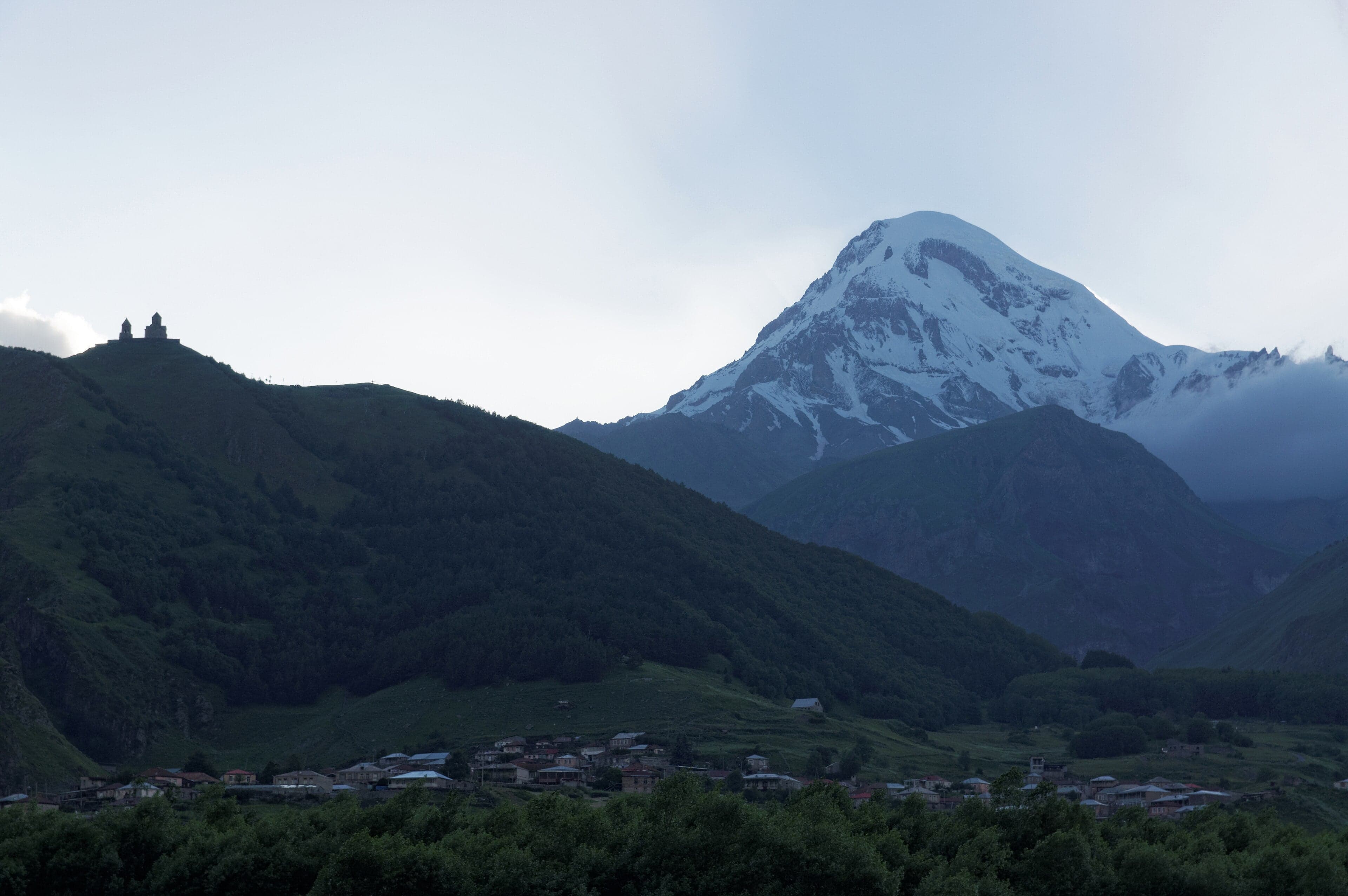 Mount Kazbek covered in snow rising above clouds with the Gergeti Trinity Church on a ridge below